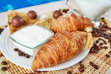 Croissants, pancakes and round chocolates are on a plate with a yogurt bowl with kefir. Below there is a mat and coffee beans. Selective focus. Continental breakfast