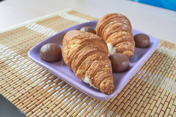 Two croissants and chocolate round candies lie on a lilac plate. Selective focus