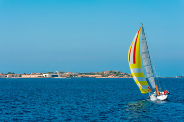 A view of a colorful lonely sailboat as it sails tilted by the wind in the Mediterranean sea with the coast in the background on a sunny day with the blue sky on summer, in Sardinia Italy
