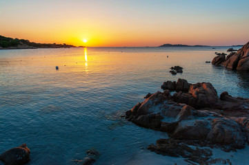 A view of a beach without people with the sun rising and brightening the rocks with dawn colors on a summer day holiday, in Sardinia Italy