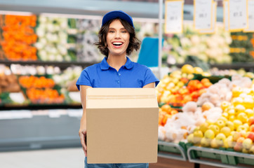 food, service and shipment concept - happy smiling delivery girl with box in blue uniform over supermarket or grocery store on background
