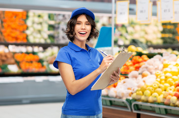 food, service and people concept - happy smiling delivery girl in blue uniform with clipboard and pen writing over supermarket or grocery store on background