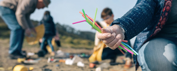 Woman hand showing handful of straws collected on the beach with group of volunteers working in the background. Selective focus in straws in foreground