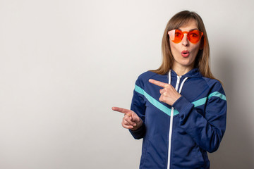 surprised young woman in a 90s sports jacket and red glasses on a light background. Model points...