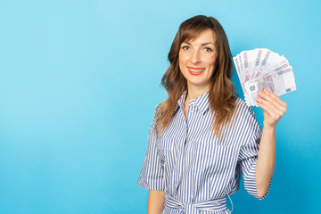 Portrait of a young friendly woman with a smile in a dress holding a fan of money on an isolated blue background. Emotional face. Concept of wealth, win, credit. Gesture of joy from winning