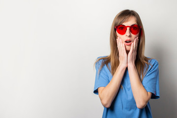 Portrait of a young friendly woman with a surprised face in a casual blue t-shirt, red glasses, holding her hands on her chin on an isolated light background. Emotional face. Gesture surprise shock