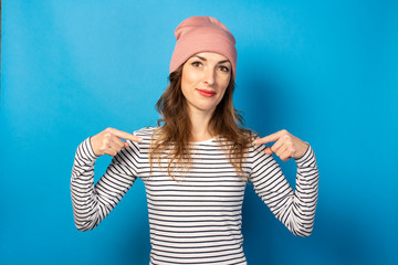 Cute young woman in a hat and striped sweater points her fingers at herself on a blue background....