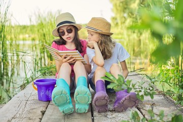 Children, two girls sitting on wooden pier in lake, talking and reading notebook