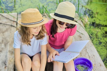 Children, two girls sitting on wooden pier in lake, talking and reading notebook