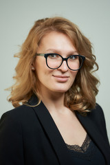 Fototapeta premium A stylish young woman in glasses dressed in a black suit with a black shirt. A close-up portrait of a happy blond lady who wears a business outfit. A concept of an office style wardrobe.