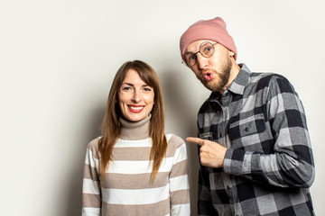 Young man with a beard in a hat and a plaid shirt with a surprised face hugs a girl in a sweater and points a finger at her on light background. Emotional face. Happy couple, meeting, a happy person