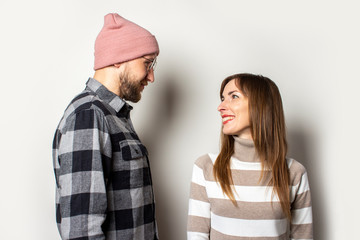 Young man with a beard in a hat and a plaid shirt and a girl in a sweater look at each other on an isolated light background. Emotional face. Happy couple dating
