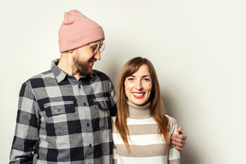 Young man with a beard in a hat and a plaid shirt hugs a girl in a sweater on an isolated light background. Emotional face. Happy couple dating