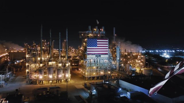 American flag waves on the wind against the background of modern petroleum refinery working at night. The plant constructions are brightly lit with technical lights of gold color. Aerial, 4K