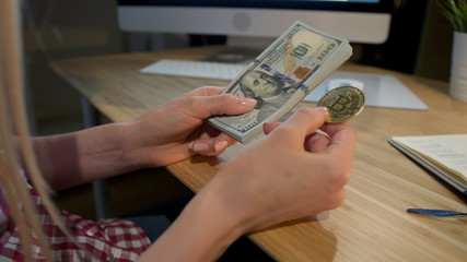 Crop view of female hands holding metal bitcoin and bundle of dollar banknotes sitting at wooden desk with notepad and computer monitor at night