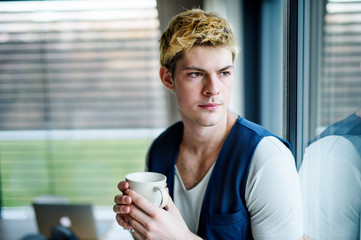 Young man with cup of coffee indoors at home, looking out of window.