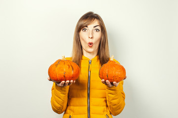 shocked young girl in a yellow jacket holds two pumpkins on a light background. Halloween concept, autumn, celebration