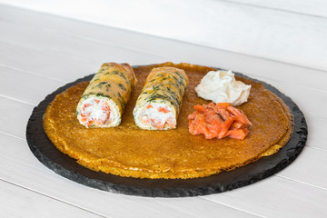 Stuffed crepes with smoked salmon, cream cheese and dill inside the dough. Thin pancakes with filling. Served on black stone plate on white wooden table. Russian fried Blini. Side view.