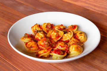 Russian traditional dish pelmeni, meat dumplings with tomato sauce, on white plat, wooden background, side view, selective focus