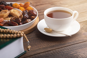 iftar concept, white Cup of tea, plate of dried fruit, Koran with prayer beads on brown wooden table in sun