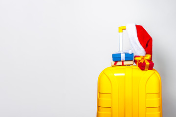 On a yellow plastic suitcase are gifts and wearing a red Santa Claus hat on a light background