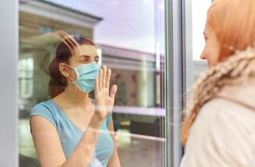 health, safety and pandemic concept - sick young woman wearing protective medical mask looking through window and waving hand to her friend outdoors