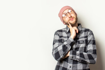 Portrait of a young man with a beard in a hat, plaid shirt and glasses looks up on an isolated light background. Emotional face. Dream gesture planning