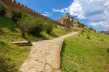 Fototapeta na wymiar An ancient fortress on top of a mountain and path along the fortification wall in sunny day. Genoese fortress, Sudak, Crimea. Historic old building. Monument of architecture. 