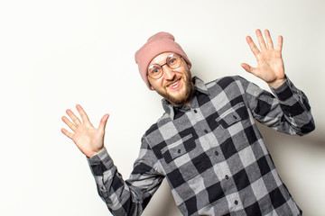 Naklejka premium Portrait of a young man with a beard in a hat, a plaid shirt peeps from behind the edge of the picture on an isolated light background. Emotional face. Gesture surprise shock