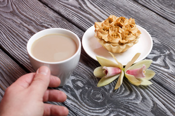 Male hand with a cup of coffee with milk. Nearby is a cake decorated with butter cream flowers. Orchid flowers are lying. On brushed pine boards painted black.