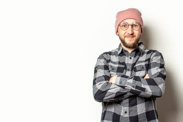 Portrait of a young man with a beard in a hat, plaid shirt and glasses with arms crossed on his chest smiling on an isolated light background. Emotional face