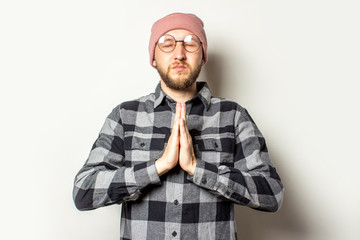 Portrait of a young man with a beard in a hat, plaid shirt and glasses is meditating or praying on...
