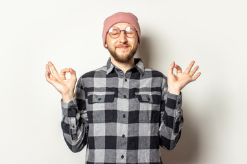 Naklejka premium Portrait of a young man with a beard in a hat, plaid shirt and glasses is meditating or praying on an isolated light background. Emotional face. Gesture of meditation, relax