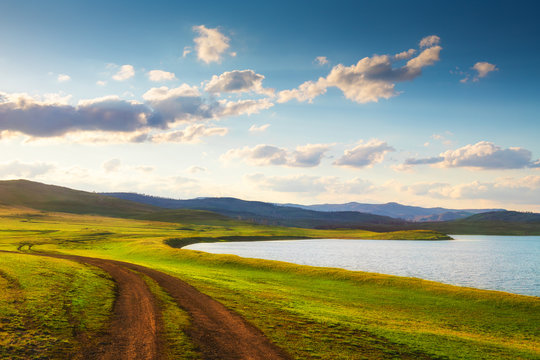 Road near the lake in the mountains. Spring nature landscape at sunset. Fresh green grass on the hills. South Ural, Bashkortostan Republic, Russia.