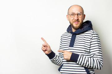 Portrait of a Bald Young man with a beard in glasses, a sweater with a hood points fingers to the side on an isolated light background. Emotional face. Gesture pay attention, look at this