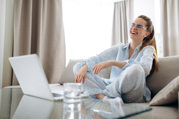 Zoom conference. Cheerful attractive girl happily communicates with friends via video calling using a laptop while sitting on a sofa in home clothes