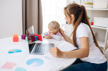 Young attractive mom works remotely on laptop at home, and her little adorable daughter is sitting nearby and watching a cartoon on a telephone. Work at home