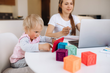 Work at home. Young attractive mom works remotely on laptop at home, and her little adorable daughter is sitting nearby and watching a cartoon on a telephone