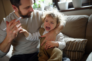 Father giving syrup to crying small sick daughter indoors at home.