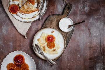 pancakes with blood oranges, nuts and cream in plates on wooden background, top view, traditional breakfast concept