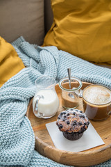 Blaubeermuffin mit Kaffee, Milch und Zucker auf Holztablett und gemütliches Sofa mit blau Wolldecke und gelb Kissen 