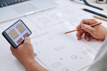 Construction engineer looking at drawing of house on smartphone screen and drawing blueprint for client