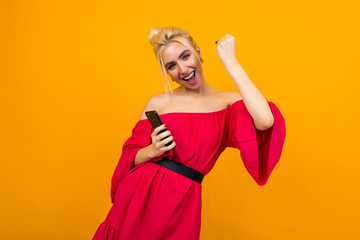 European blond girl in a red dress smiles with joy holding a phone in her hands on a yellow studio background with copy space