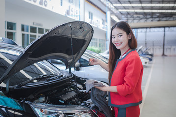woman near car's hood. Beautiful Asian girl in covered parking of shopping center, stands near car with raised engine compartment hood, checks engine oil level in engine, inspects feeler gauge - obrazy, fototapety, plakaty