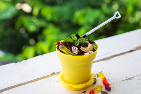 High Angle View A Cookie Crumbs In A Yellow Pot Decorated With Worm Jelly, Chocolate Gravel And Pepermint With Toy Shovel On White Wooden Table With Green Nature Background