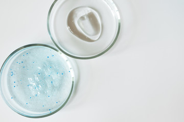 Blue shower gel with scrub grain with bubbles and transparent shampoo smudge in glass petri dish on white background. Concept laboratory tests and research, making cosmetic