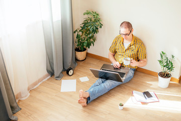 Young man is siiting on the floor and working at home on a computer with the phone, papers and a cup of coffee. Remote work, work from home.