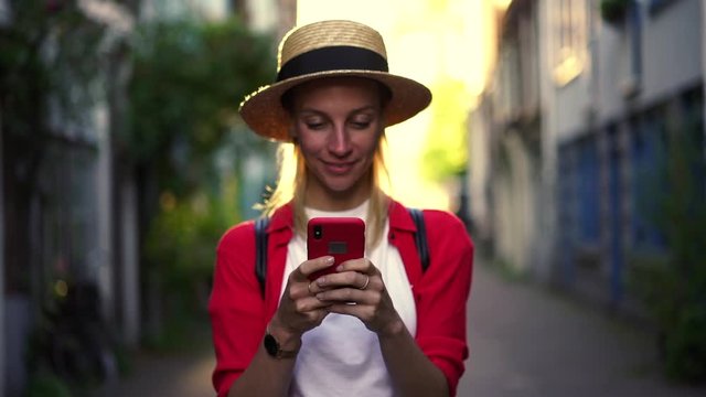 Smiling female tourist enjoying writing messages to friend using 4g internet on mobile phone, happy millennial hipster girl in hat feeling excited while chatting online on smartphone. Booking apps

