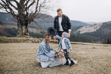 Fototapeta premium Stylish couple in the mountains. Stock Photo. A guy in a leather jacket and a young girl in a gray-blue wedding dress and a denim jacket with their son on the background of a large tree and sky