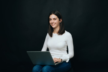 Smiling girl is enjoying working on her computer while sitting on a chair on black background.